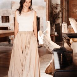 Smiling woman in a white sleeveless top and long beige skirt stands beside a piano in a rustic, wooden interior.