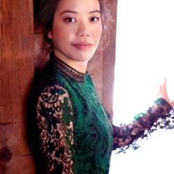 Portrait of an Asian woman in a green lace dress standing beside a wooden beam, sunlight on the right side.