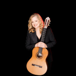 Smiling woman in a black blazer holding an acoustic guitar against a black background.