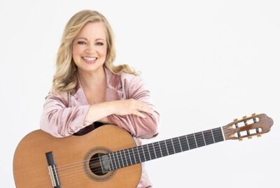 Smiling woman in a pink jacket holds an acoustic guitar across her torso, posing for a portrait.