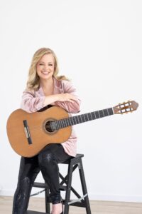 Smiling woman sits on a stool, cradling an acoustic guitar across her lap, wearing a pink velvet blazer.