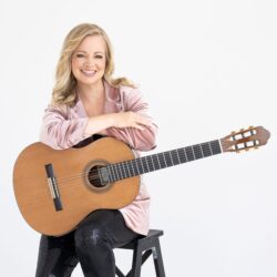 Smiling woman sits on a stool, cradling an acoustic guitar across her lap, wearing a pink velvet blazer.